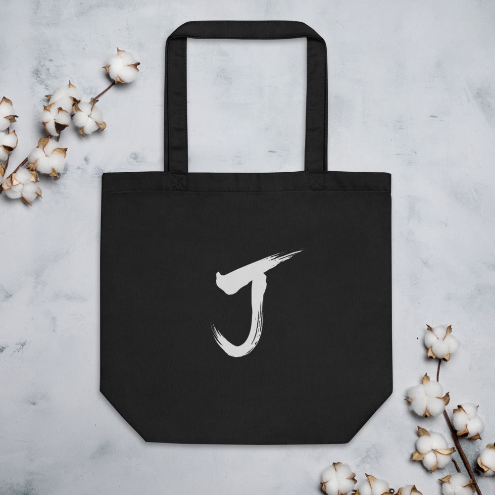 The letter J rendered in a single black sumi-e brush gesture, printed on a canvas tote bag. Flat lay, Ink colourway.