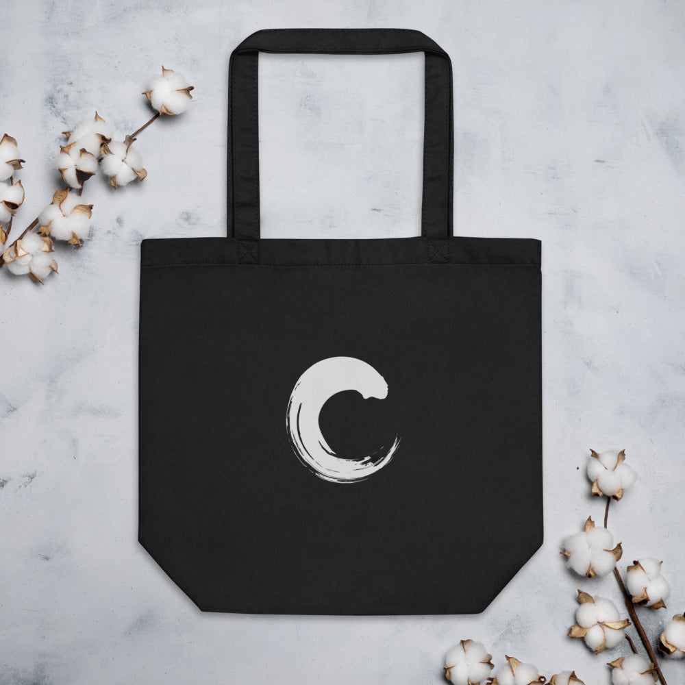 The capital letter C printed as a black sumi-e brush gesture on a tote bag. Flat lay, Ink colourway.