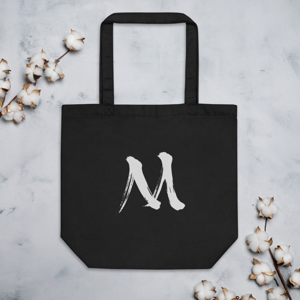 The letter M in black sumi-e brush stroke style printed on a minimalist tote bag. Flat lay, Ink colourway.