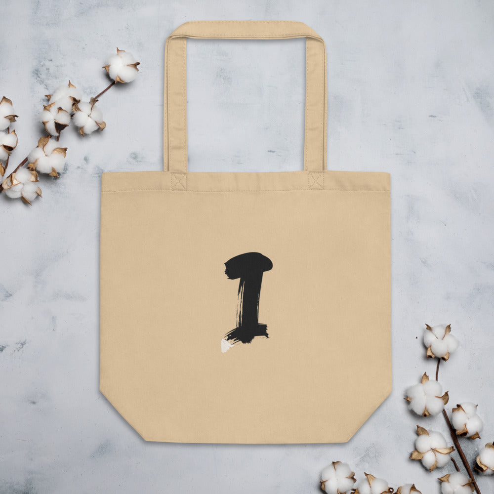 The letter I in a single vertical sumi-e brush gesture, printed on a canvas tote bag. Flat lay, Bone colourway.