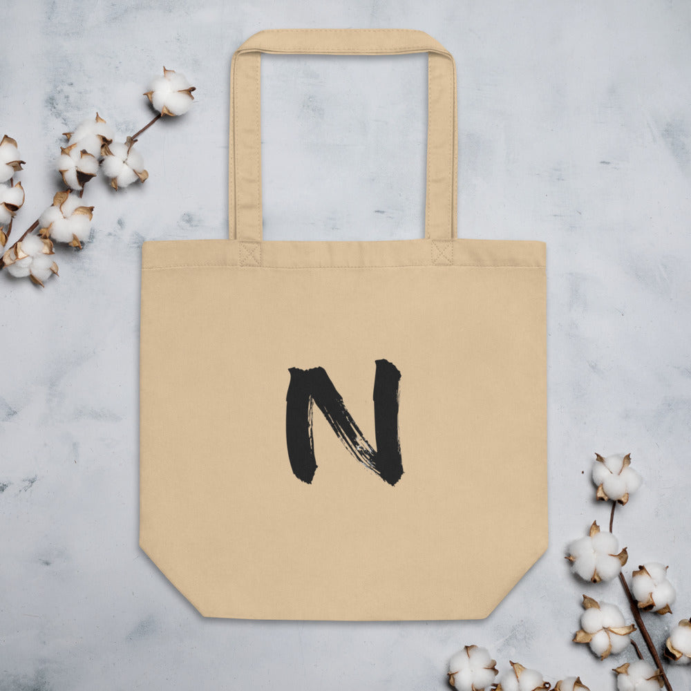A minimal black sumi-e ink brush stroke of the letter N printed on a canvas tote bag. Flat lay, Bone colourway.