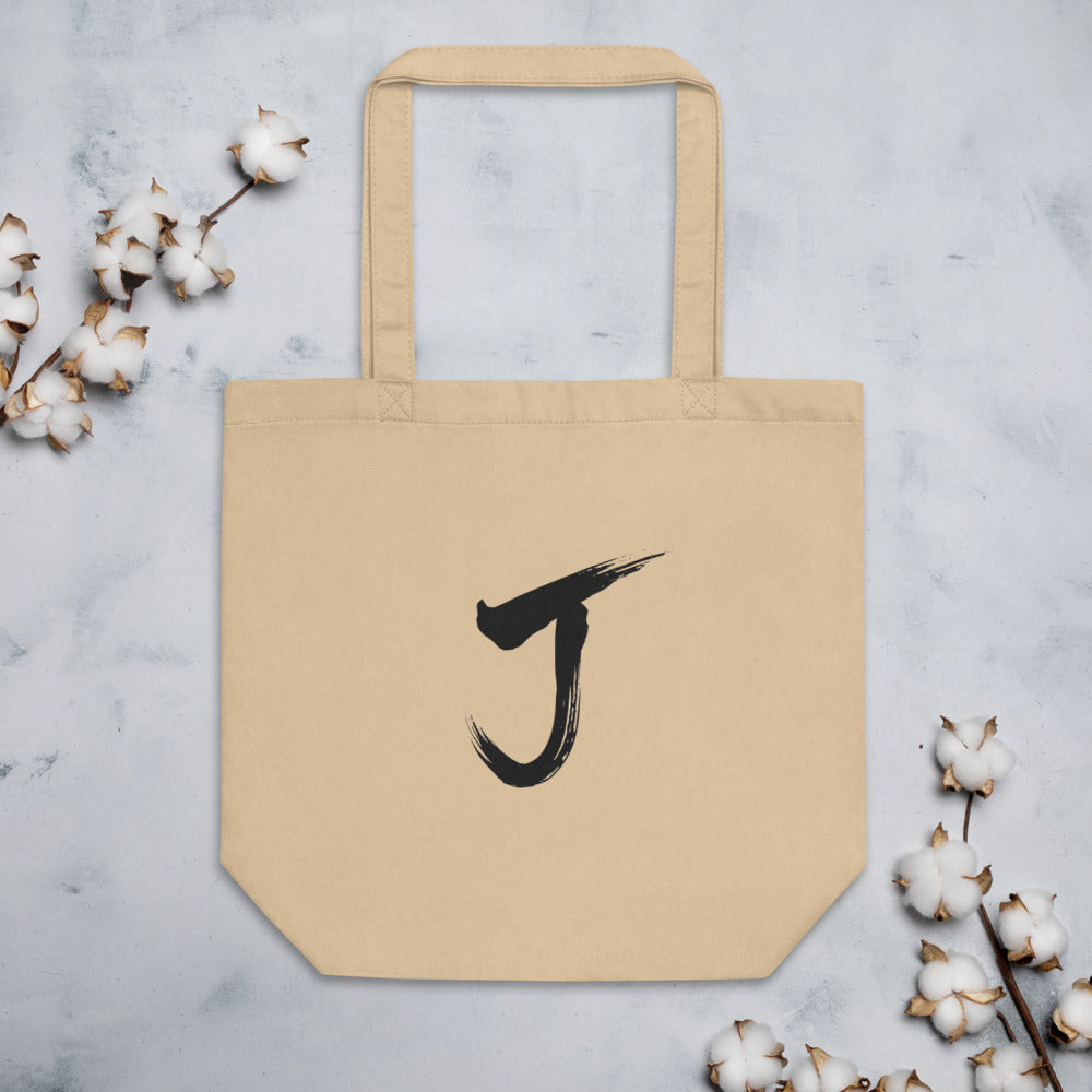 The letter J rendered in a single black sumi-e brush gesture, printed on a canvas tote bag. Flat lay, Bone colourway.