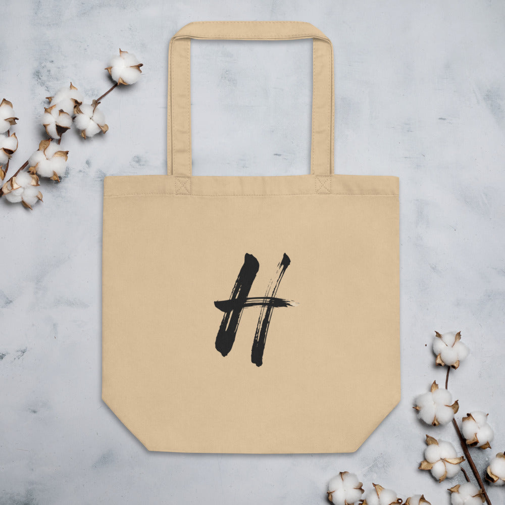 The capital letter H rendered in black sumi-e brushstrokes printed on a tote bag. Flat lay, Bone colourway.
