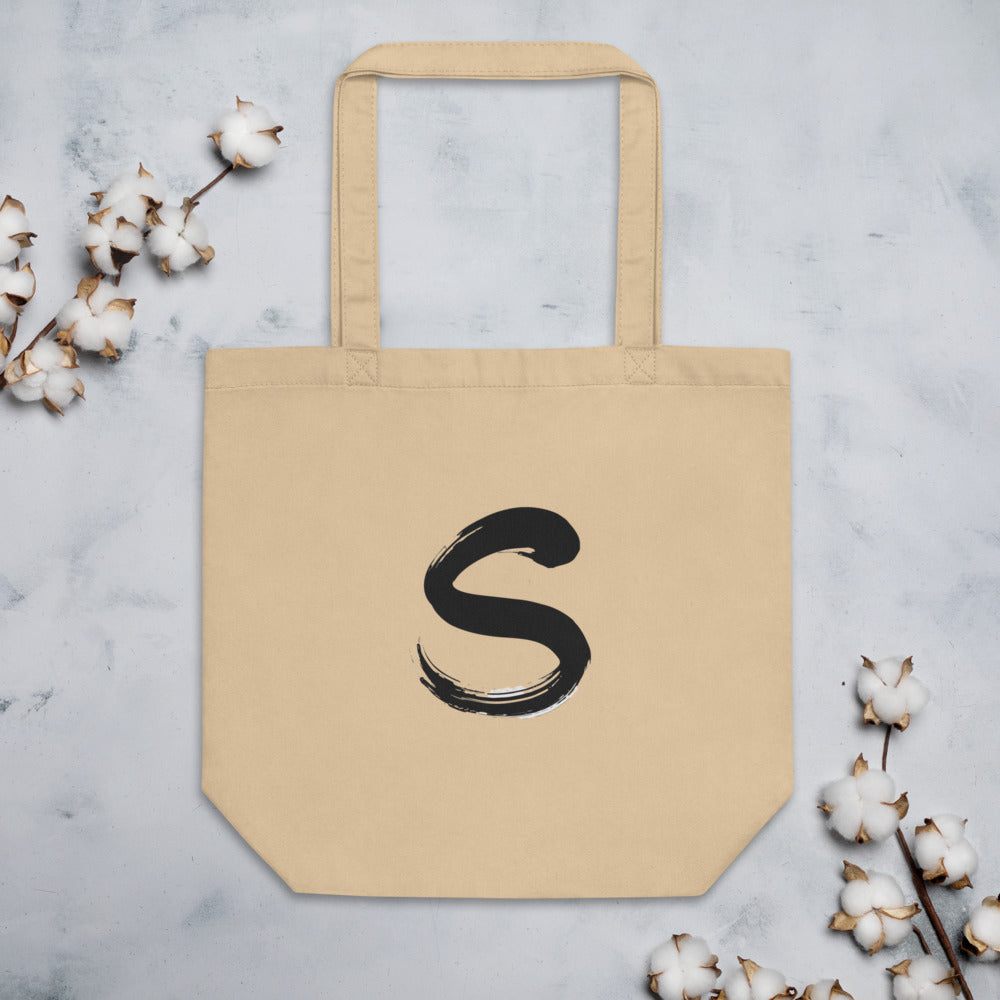 The letter S rendered in a single serpentine sumi-e brush stroke printed on a tote bag. Flat lay, Bone colourway.
