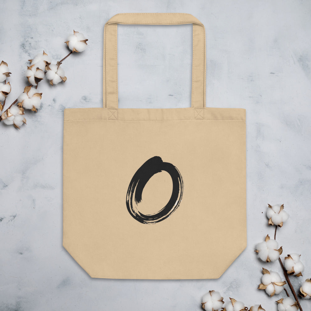 A single black sumi-e brushstroke forming an unclosed letter O on a tote bag. Flat lay, Bone colourway.