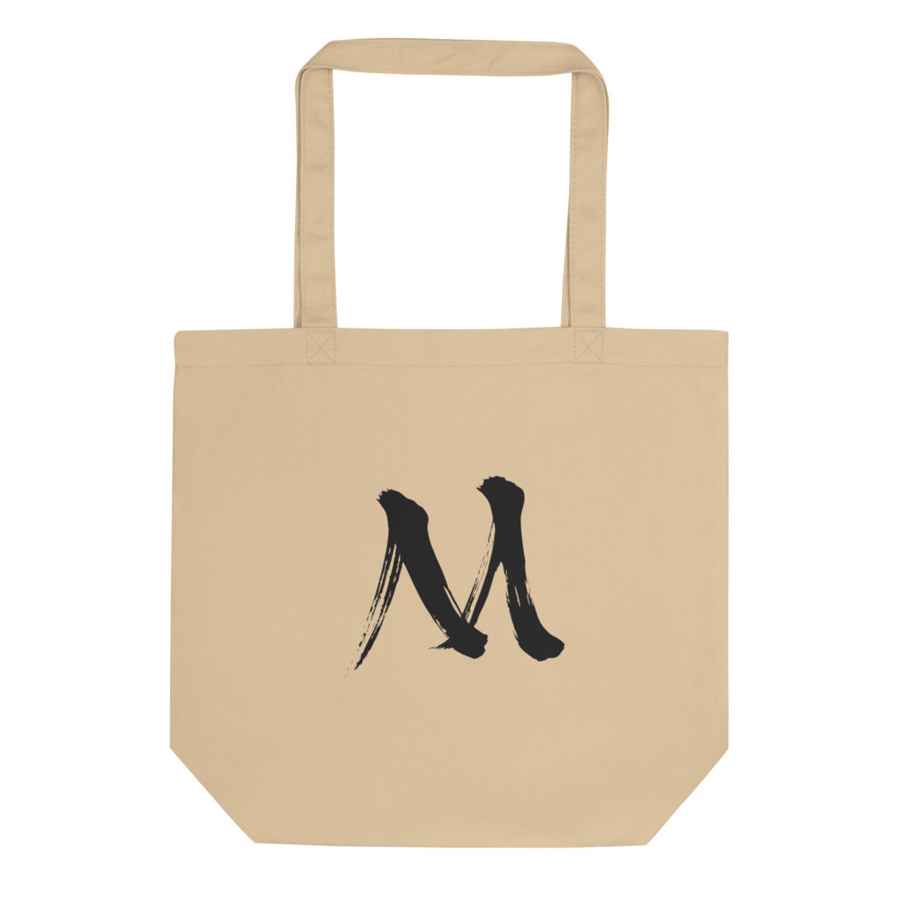 The letter M in black sumi-e brush stroke style printed on a minimalist tote bag. Shown on the Bone (Oyster) colorway.