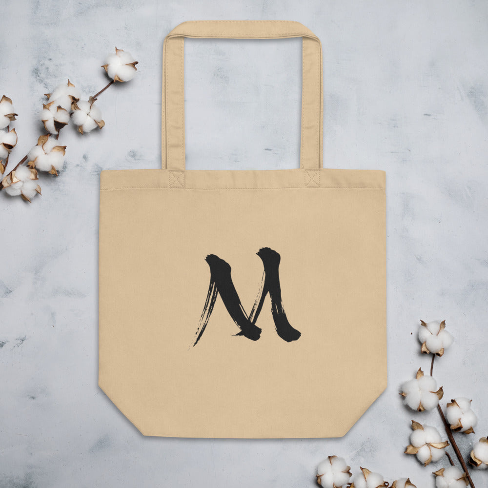 The letter M in black sumi-e brush stroke style printed on a minimalist tote bag. Flat lay, Bone colourway.