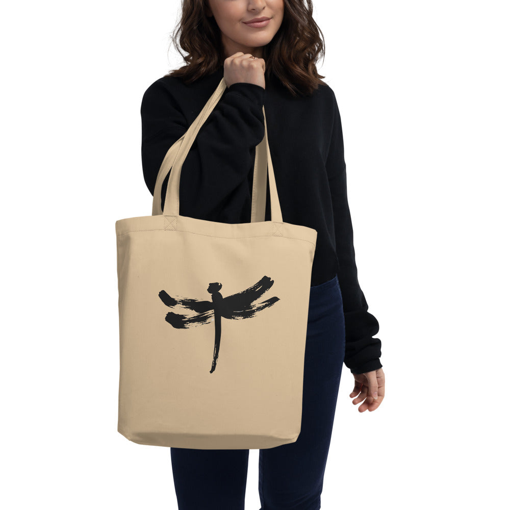 A minimalist dragonfly sumi-e ink design printed on a canvas tote bag. Carried over the shoulder, Bone colourway.