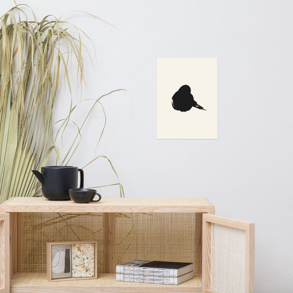 A minimalist resting sparrow printed in black sumi-e ink on an art print, showing a single wet brushstroke body. Above a rattan cabinet, 12″×16″ size.